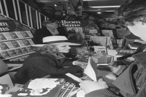 Eine Frau mit Hut signiert Autogramme für einen Mann auf einer Buchsignierveranstaltung, umgeben von einer Gruppe von Menschen, einige halten Bücher, mit Bücherregalen voller Bücher im Hintergrund, in Schwarz-Weiß, mit einem Geländer auf der linken Seite.