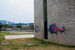 Betongebäude mit Graffiti an der Seite, umgeben von Gras, Bäumen, Strommasten, Fahrzeugen auf der Straße, anderen Gebäuden, Hügeln und einem klaren blauen Himmel.