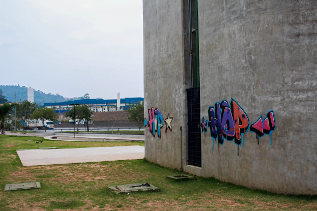 Betongebäude mit Graffiti an der Seite, umgeben von Gras, Bäumen, Strommasten, Fahrzeugen auf der Straße, anderen Gebäuden, Hügeln und einem klaren blauen Himmel.
