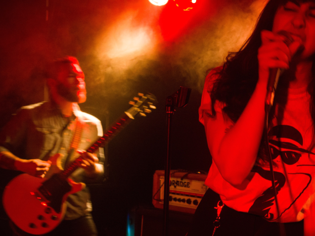 Eine Frau singt in ein Mikrofon auf der Bühne, während ein Mann eine akustische Gitarre im Hintergrund spielt und Deckenlampen die lebendige Szenerie beleuchten.