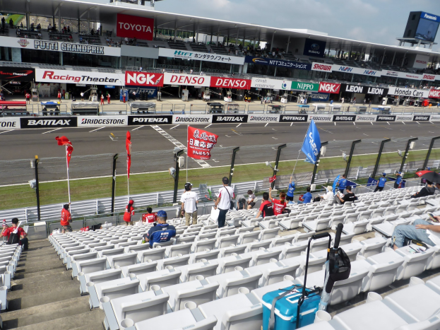 Eine Gruppe von Menschen auf Tribünen bei einer Rennstrecke mit Fahnen, Schildern, Geländern und Stufen, mit einem Stadion und einem bewölkten Himmel im Hintergrund.