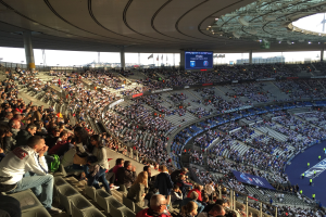 Eine große Menschenmenge in einem Stadion schaut ein Fußballspiel zu, mit einer Bühne rechts, Fahnen, Stangen, einem Bildschirm und der Allianz Arena in München im Hintergrund.