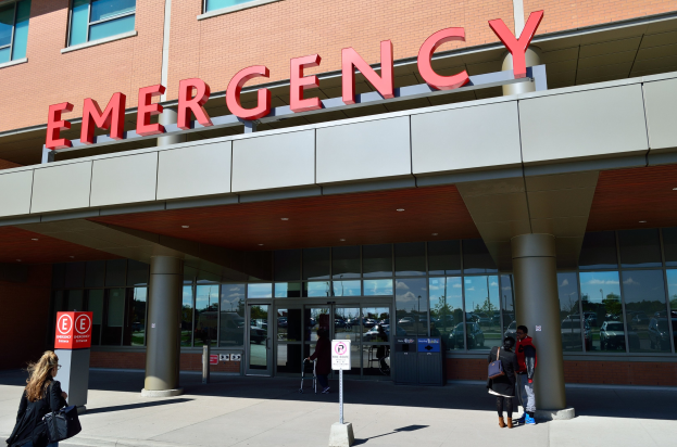 Gruppe von Menschen, die auf ein Notaufnahme-Gebäude mit Glaswänden, Säulen, einem Namensschild und umliegenden Schildern und einem Mülleimer zugehen.