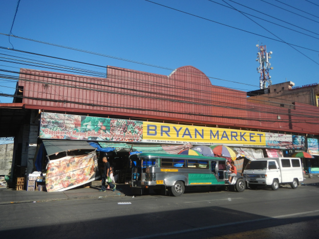Eine belebte Straßenszene mit Fahrzeugen, Fußgängern, Ständen, Gebäuden und einem geparkten Bus vor einem Gebäude mit einer "Bryan Market"-Aufschrift.