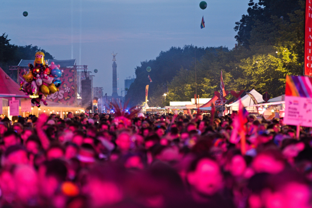 Große Menschenmenge auf einem nächtlichen Musikfestival mit bunten Lichtern, Ballons, Zelten, Bannern, Flaggen, Bäumen, einem Turm und einem sternenklaren Himmel im Hintergrund.