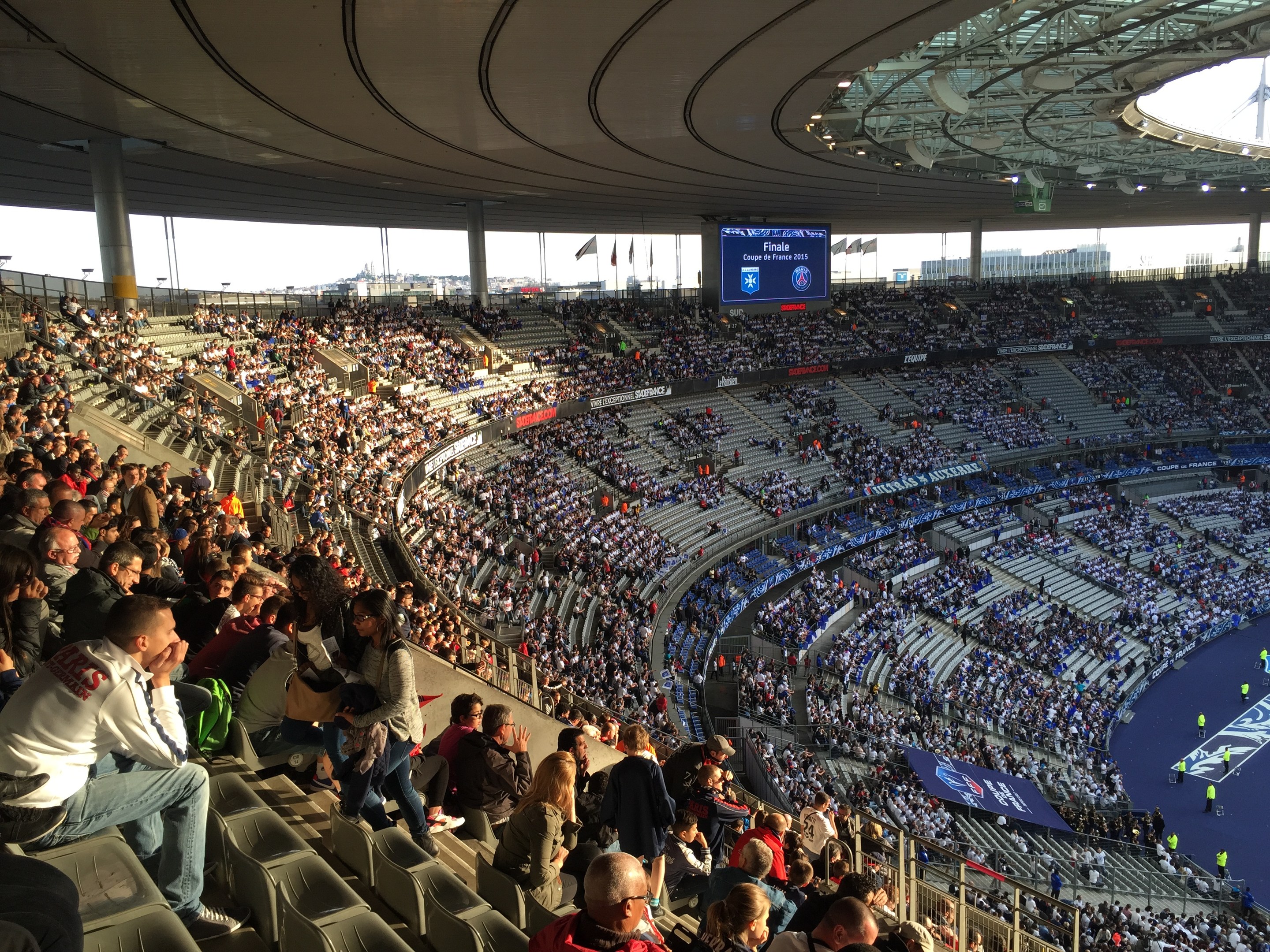 Große Menschenmenge in einem Stadion bei einem Fußballspiel, mit einer Bühne, Fahnen, Stangen, einem Bildschirm und der Allianz Arena in München, Deutschland im Hintergrund.