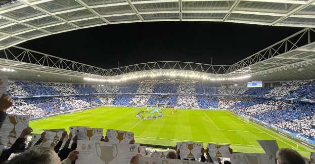 Eine Gruppe von Menschen hält Schilder vor einem Fußballstadion, einige liegen auf dem Boden, ein Bildschirm rechts und Zuschauer im beleuchteten Stadion im Hintergrund bei einer feierlichen Nachtveranstaltung.