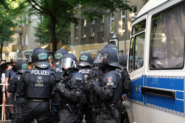 Gruppe von Polizeibeamten vor einem Bus auf einer Straße mit einer Menge hinter Absperrungen, Bäumen und Gebäuden mit Fenstern im Hintergrund.