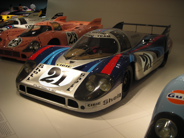 Ein Porsche 917K Le Mans Rennauto in einem Museum neben anderen Sportwagen ausgestellt, mit Menschen in der Nähe und einer Wand im Hintergrund.