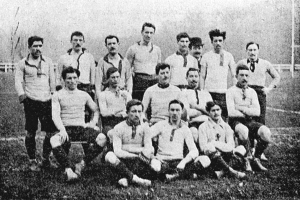 Ein Schwarz-Weiß-Foto einer Fussballmannschaft aus den 1920er Jahren, mit einigen Spielern, die stehen, und anderen, die auf dem Boden sitzen, vor einer Kulisse aus Pfählen, Bäumen und einem klaren Himmel.