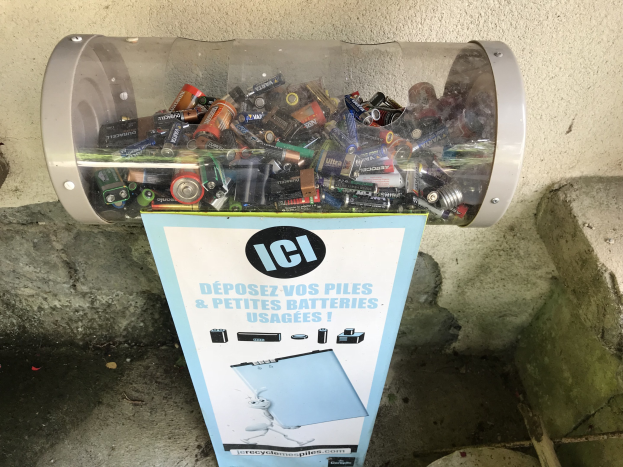 Mülltonne quillt über von benutzten Batterien auf einem Gehweg, mit einem Plakat an der Wand dahinter und Stufen rechts.