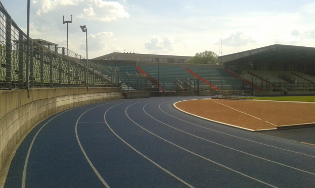 Großes Stadion mit einer zentralen Laufbahn, umgeben von Gras, Zaun, Beleuchtungsmasten, Sitzplätzen, einem kleinen Schuppen, einem Gebäude, Bäumen und einem bewölkten Himmel.