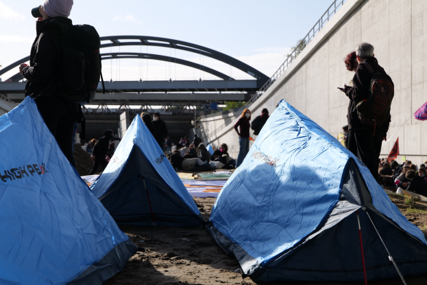 Gruppe von Menschen, die auf einem sandigen Strand in der Nähe von Zelten sitzen, mit einer Wand und einer Brücke im Hintergrund, die Gegenstände während einer Klimawandelprotest halten.