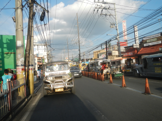 Eine belebte Stadtstraße mit Fahrzeugen, Absperrgittern, Fußgängern auf dem Gehweg, Stromleitungen, Gebäuden mit Schildern und einem bewölkten Himmel.