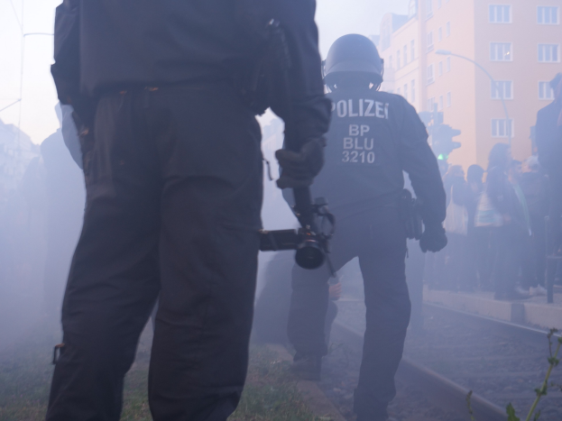 Zwei Polizeibeamte auf einem Bahngleis mit einer Gruppe von Menschen und Kameras im Hintergrund, städtische Gebäude und ein bewölkter Himmel sind zu sehen.