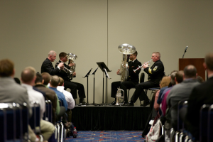 Eine Gruppe von Männern, die auf einer Bühne Blasinstrumente spielen, mit Notenständern, einem Podium mit Mikrofon und einem Publikum, das auf Stühlen sitzt.