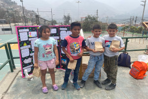 Eine Gruppe von Kindern steht auf einem Basketballfeld, jeder hält eine Papiertüte in der Hand, mit Vorräten und einer Texttafel in der Nähe und einer Kulisse aus Zäunen, Pfosten, Bäumen, Gebäuden und Hügeln.