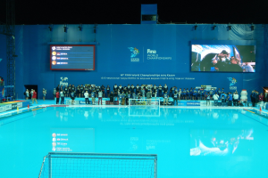 Eine Gruppe von Menschen um ein Hallenbad mit Netz am Boden herumstehend, mit einer Wand im Hintergrund, die den Text und Logos "FINA World Championships 2015 Kazan" anzeigt, sowie Gerüste und Beleuchtung.