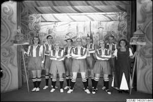 Eine Gruppe strahlender Frauen in einem 1940er-Soccer-Team posiert auf einer Bühne, jede hält einen Stock in der Hand, mit einer bemalten Wand im Hintergrund.