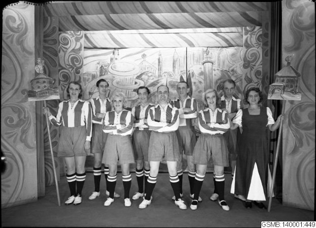 Eine Gruppe strahlender Frauen in einem 1940er-Soccer-Team posiert auf einer Bühne, jede hält einen Stock in der Hand, mit einer bemalten Wand im Hintergrund.