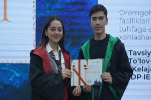 Ein Mann und eine Frau in akademischen Roben stehen nebeneinander, jeder hält ein Zertifikat in der Hand, mit einem Bildschirm im Hintergrund.