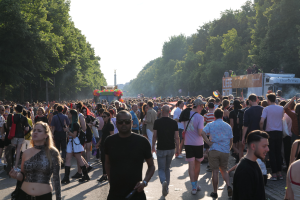 Eine große Menschenmenge, die eine von Bäumen gesäumte Straße entlanggeht, mit einem Turm im Hintergrund und Fahrzeugen mit Menschen auf der rechten Seite, wahrscheinlich beim Christopher Street Day in Berlin unter einem klaren blauen Himmel.