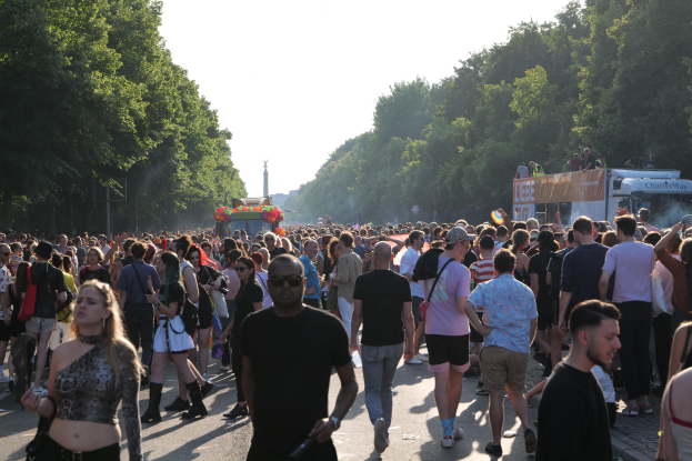 Eine große Menschenmenge, die eine von Bäumen gesäumte Straße entlanggeht, mit einem Turm im Hintergrund und Fahrzeugen mit Menschen auf der rechten Seite, wahrscheinlich beim Christopher Street Day in Berlin unter einem klaren blauen Himmel.