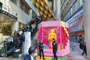 Eine Gruppe von Menschen steht vor einem Glaswand-Gebäude mit Texttafeln, einige tragen Masken, in der Nähe einer Rolltreppe; eine pinkfarbene Box in der Mitte zeigt Schaufensterpuppen und trägt 'Netflix's new Netflix booth in Tokyo.'