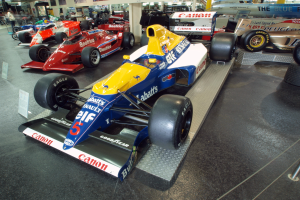 Eine Gruppe von Rennwagen, die in einem Museum ausgestellt sind, mit Besuchern, die sie zwischen Tafeln, Säulen, Lampen und anderen Ausstellungsstücken bewundern.