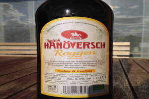 Flasche Bier mit Etikett auf einem Holztisch, mit einem Glasfenster im Hintergrund, das ein Gebäude und den Himmel zeigt.