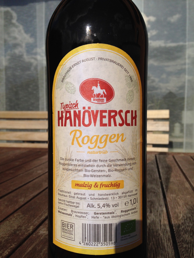 Flasche Bier mit Etikett auf einem Holztisch, mit einem Glasfenster im Hintergrund, das ein Gebäude und den Himmel zeigt.