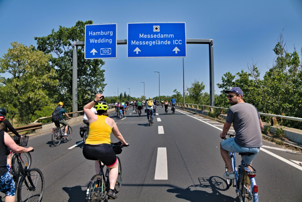 Gruppe von Radfahrern in Helmen auf einer Straße mit einer Begrenzung und Bäumen auf beiden Seiten, Laternen im Hintergrund, unter einem klaren blauen Himmel, mit einem Schild, das eine Radtour in Hamburg anzeigt.