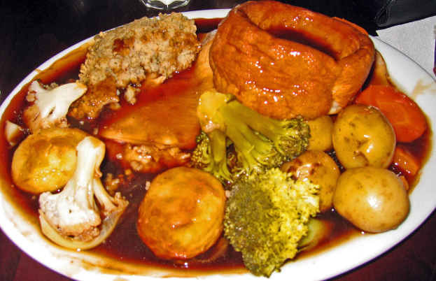 Ein weißer Teller mit Yorkshire-Pudding umgeben von Karotten, Kartoffeln und Zwiebeln auf einem Tisch mit Gläsern und anderen Gegenständen.