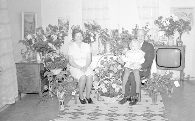 Schwarzes und weißes Foto einer Familie aus drei Personen - einem Mann, einer Frau und einem Kind - auf einem Sofa sitzend mit Blumenvasen auf den Seitentischen, einem Teppich auf dem Boden, gerahmten Wanddekorationen und Vorhängen im Hintergrund.