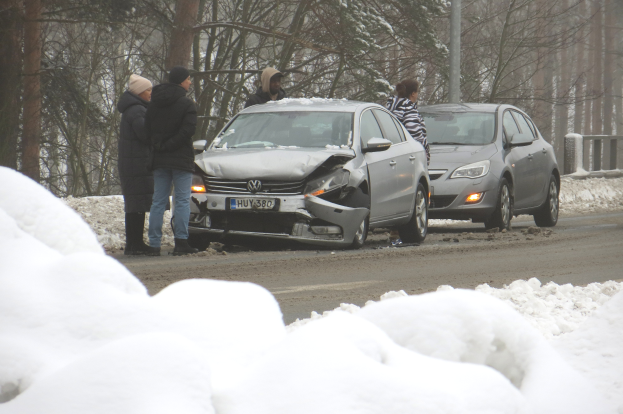 Ein verunglücktes Auto am Rande einer verschneiten Straße, umgeben von Menschen, mit Bäumen und einem Mast im Hintergrund.