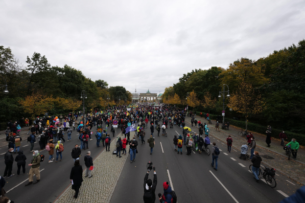 Eine große Gruppe von Menschen marschiert auf einer von Bäumen gesäumten Straße in Berlin während einer Protestdemonstration, wobei einige Kameras halten und ein Gebäude im Hintergrund zu sehen ist.