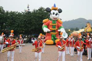 Eine Gruppe von Menschen in Kostümen spielt Musikinstrumente in der Nähe eines Mickey-Maus-Floats während eines Umzugs, mit Bäumen, Gebäuden und einem klaren Himmel im Hintergrund; ein Schild mit Text ist auf der linken Seite sichtbar.