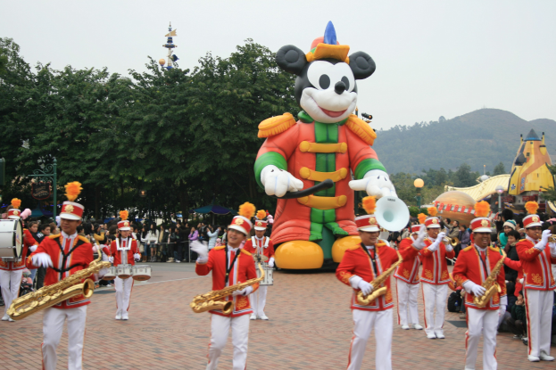 Eine Gruppe von Menschen in Kostümen spielt Musikinstrumente in der Nähe eines Mickey-Maus-Floats während eines Umzugs, mit Bäumen, Gebäuden und einem klaren Himmel im Hintergrund; ein Schild mit Text ist auf der linken Seite sichtbar.