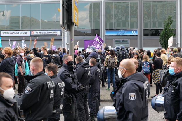 Eine große Gruppe von Menschen steht vor einem Gebäude, einige halten Schilder und tragen Helme, im Vordergrund ein Pfahl mit einer Schilder und im Hintergrund ein Baum, was auf eine Demonstration hindeutet.