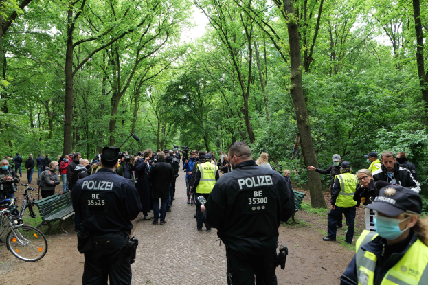 Gruppe von Polizisten vor einer Menschenmenge bei einer Outdoor-Veranstaltung mit Fahrrädern und einer Bank im Vordergrund, Bäume und Himmel im Hintergrund.