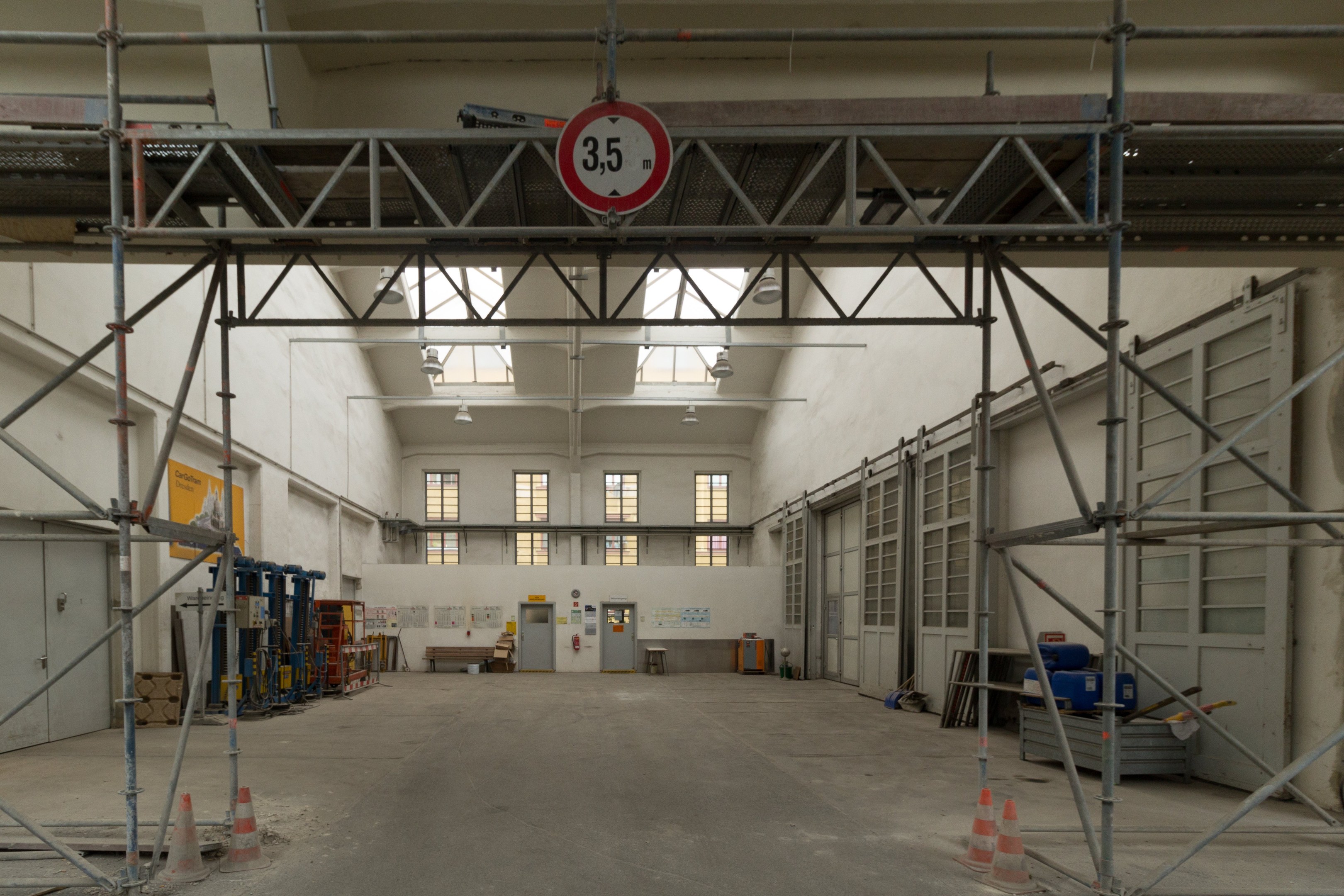 Lagerhalle mit Gerüst, Verkehrskegeln, Metallstangen, einem Schild, Maschinen, einer Tür, Fenstern, Wänden, Deckenlampen und einem Dach.