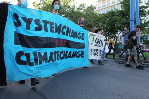 Eine Gruppe von Menschen marschiert mit einem "System Change Not Climate Change"-Schild die Straße entlang, einige tragen Masken und Fahrräder stehen in der Nähe, unter einem klaren blauen Himmel mit Bäumen und Gebäuden im Hintergrund.