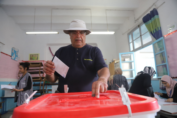 Ein Mann mit Hut stimmt in einer Wahllokal ab, vor einer roten Wahlurne stehend, mit einem Stimmzettel in der Hand, während andere an Tischen sitzen und schreiben, im Hintergrund ein Fenster mit einem Vorhang.