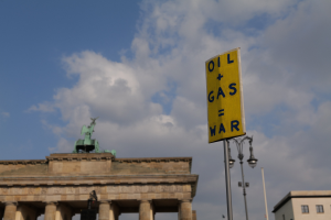Das Brandenburgische Tor in Berlin, Deutschland, mit einem gelben Schild, auf dem "Öl- und Gaskrieg" steht, im Vordergrund vor einem bewölkten Himmel.