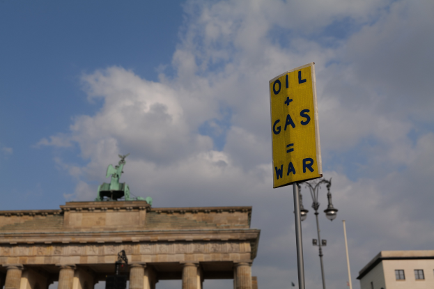 Das Brandenburgische Tor in Berlin, Deutschland, mit einem gelben Schild, auf dem "Öl- und Gaskrieg" steht, im Vordergrund vor einem bewölkten Himmel.