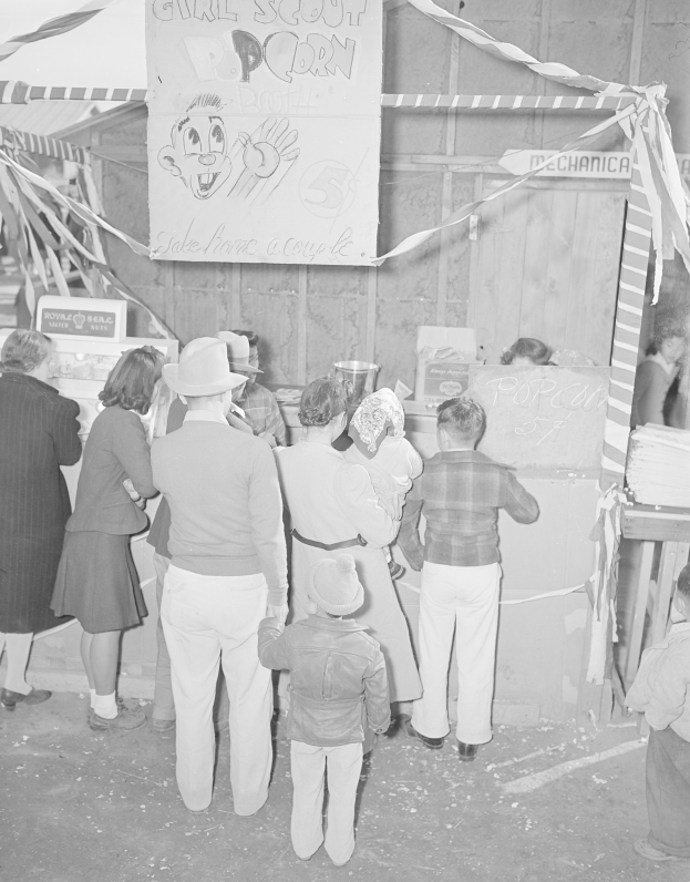 Schwarzes und weißes Bild von Menschen, die vor einer Karnevalsbude mit Bändern und einer Schautafel stehen.