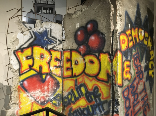 Mauer mit Graffiti, das "Freiheit und Demokratie" in fetten schwarzen Buchstaben liest, ein Geländer unten und ein Gebäude mit Fenstern im Hintergrund.