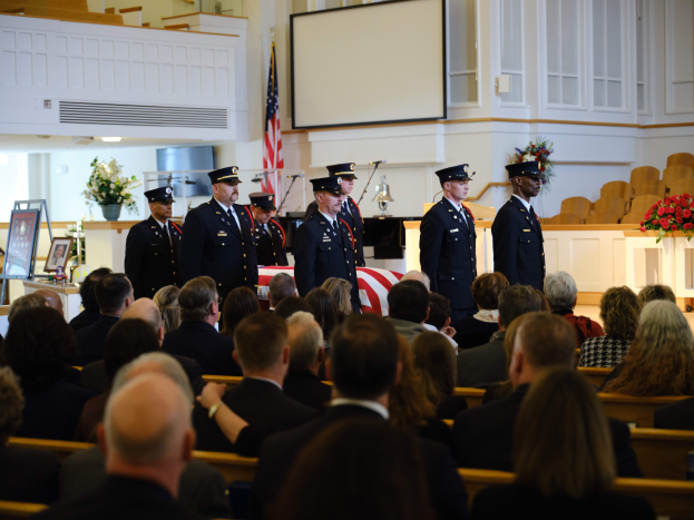 Eine Gruppe uniformierter Personen mit Mützen, die vor sitzenden Trauergästen an einer Beerdigungszeremonie stehen, mit Blumensträußen, einer Fahne, einem Bildschirm und einer Wand im Hintergrund.