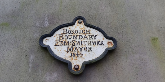 Ein Metallschild an einem Gebäude mit schwarzem Hintergrund und weißer, fetter, zentrierter Schrift, die 'Borough Boundary Edm Smithwick Mayor' liest.