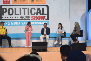 Eine Gruppe von Menschen sitzt auf einer Bühne mit einer Frau, die ein Mikrofon in der Mitte hält, einem Publikum im Vordergrund und einem Banner mit der Aufschrift "Politische Straßen-show 2019" im Hintergrund.